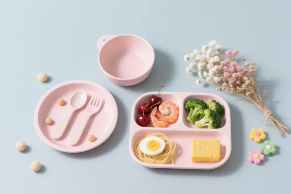 Pastel pink silicone baby feeding set with divided plate, bowl, fork and spoon, styled with food and flowers