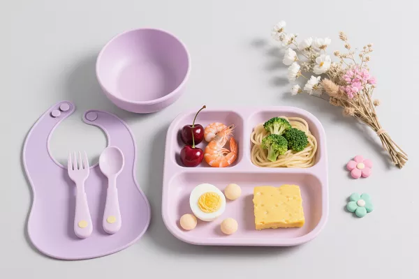 Lavender silicone baby feeding set with bib, bowl, spoon, fork, and divided suction plate, styled with food and flowers