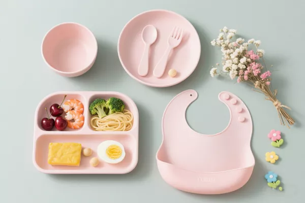 Pink silicone baby feeding set with bib, suction bowl, divided plate, spoon and fork on pastel background