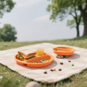 Orange silicone baby plate used for picnic with sandwich, juice, and snacks on blanket outdoors