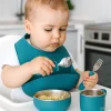 Toddler using eco-friendly teal silicone feeding set. Baby eating with teal silicone bowl, cup, spoon, and fork.