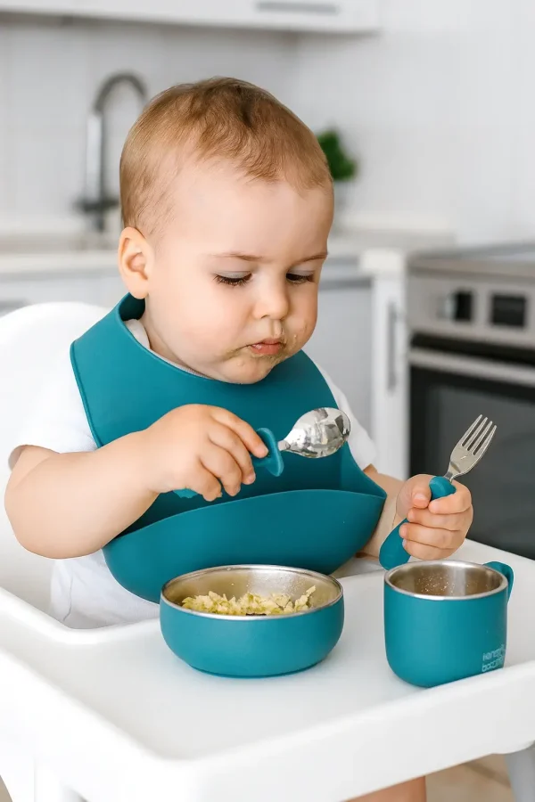 Toddler using eco-friendly teal silicone feeding set. Baby eating with teal silicone bowl, cup, spoon, and fork.